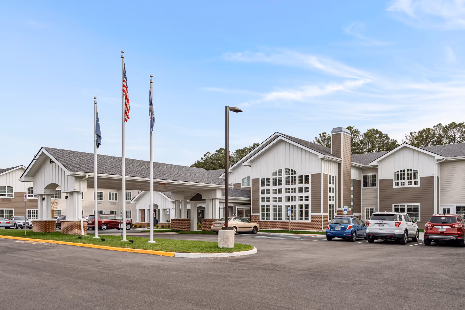 Front exterior of a senior living facility with a covered entrance, three flagpoles, large windows and parked cars.
