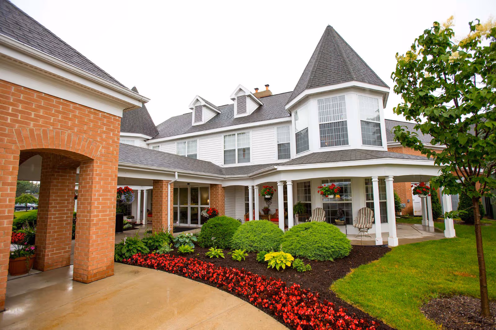 Exterior view of Independence Village Of Midland showing a large building with white siding and brick accents, a covered porch with white columns, well-maintained landscaping with green bushes and red flowers, and a tree on the right side.
