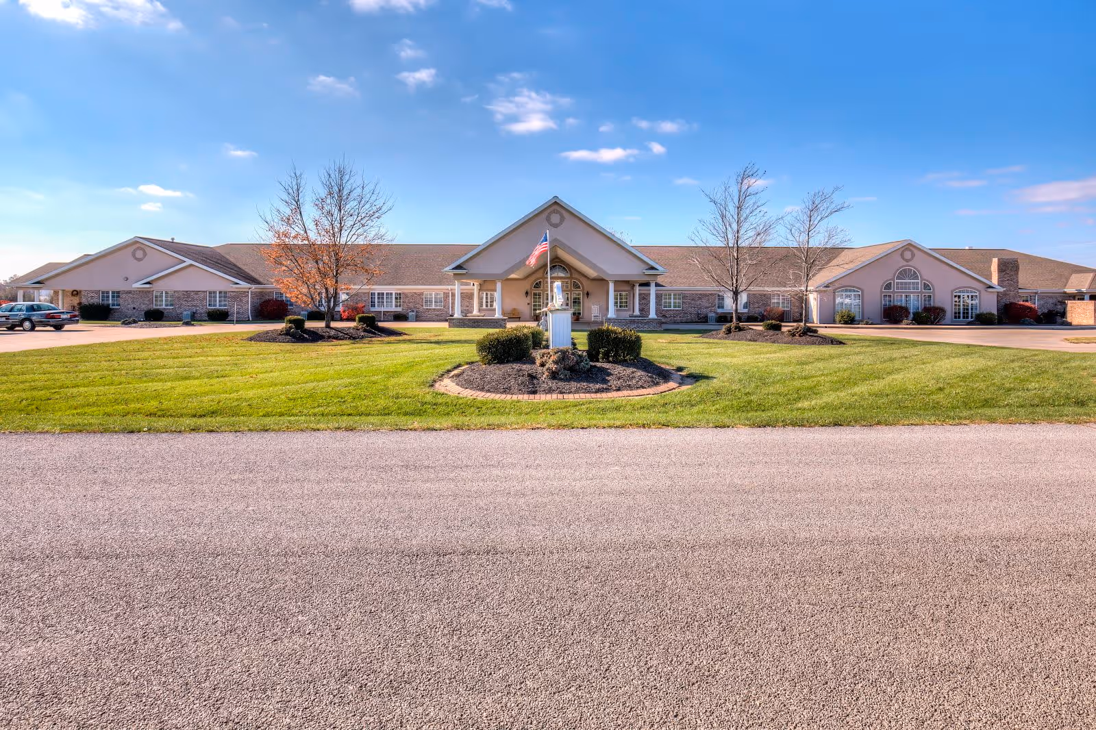 Front exterior view of a single-story senior living facility building with a well-maintained lawn, a circular landscaped area with bushes and a flagpole, and a clear blue sky.