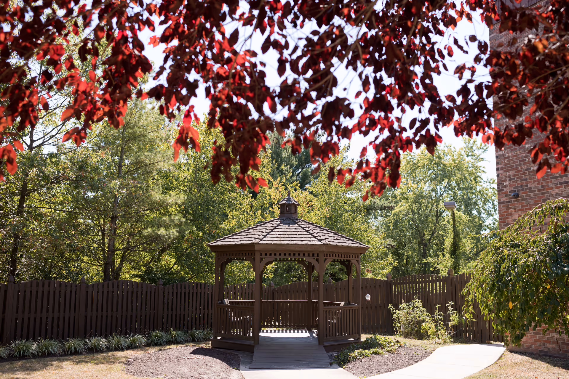 A wooden gazebo in a garden area surrounded by trees and plants, with red leaves hanging from a tree in the foreground and a brick building partially visible on the right.