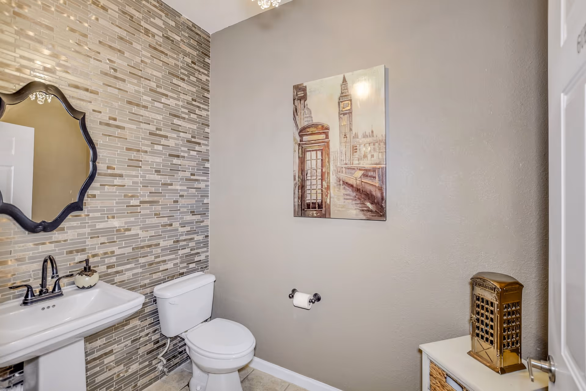 Small modern bathroom with a pedestal sink, toilet, mosaic tiled accent wall, decorative mirror, and wall art.
