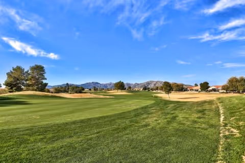A lush green golf course fairway with trees, hills, and a bright blue sky.
