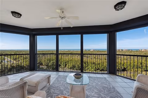 Screened-in balcony with wicker furniture including chairs and an ottoman, a glass coffee table on a patterned rug, ceiling fan above, and a view of a green forested area extending to the horizon under a clear blue sky.