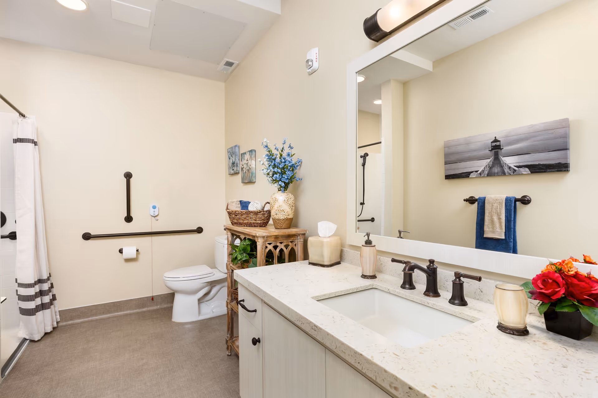 A clean and well-lit bathroom with a white toilet, a shower with a white curtain, and a countertop with a sink and faucet. The walls are painted beige, and there are decorative items including a vase with blue flowers, a basket, and a small plant on a wooden shelf next to the toilet. A large mirror is mounted above the sink, reflecting part of the room. There is a towel rack with a blue and beige towel and a black and white photo of a lighthouse on the wall.