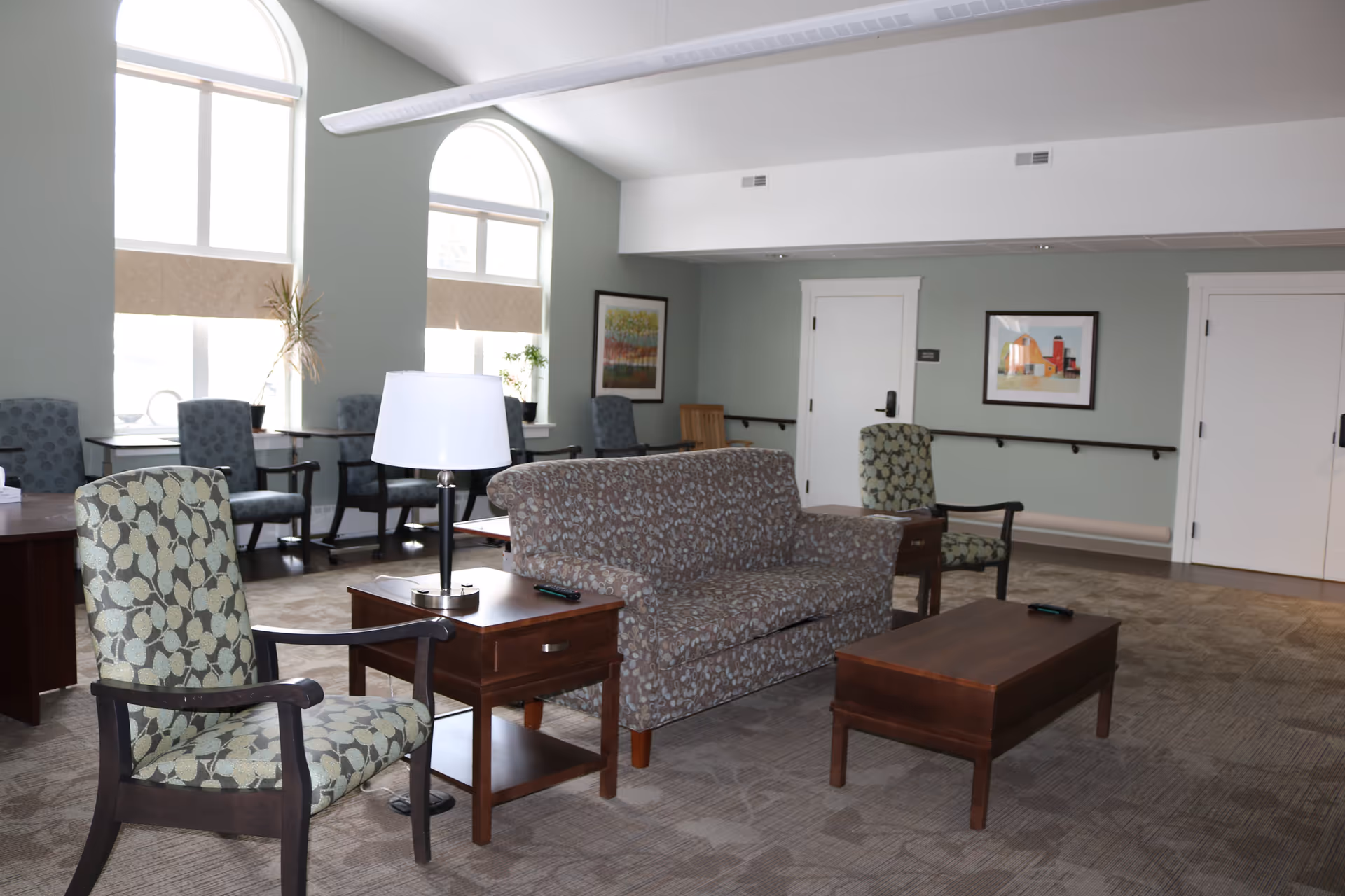A well-lit common area with patterned armchairs, a floral-patterned sofa, wooden side tables with lamps, and a wooden coffee table. The room has large arched windows with blinds, framed artwork on the walls, and a carpeted floor.