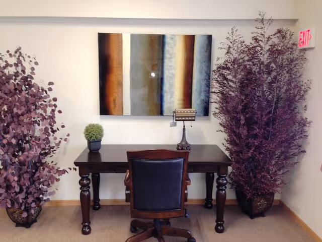 A small office or workspace with a dark wooden desk and a black leather office chair. On the desk, there is a small green potted plant and a vintage-style desk lamp. Behind the desk, a large abstract painting with vertical stripes in shades of brown, gray, and blue hangs on the wall. On either side of the desk, there are tall decorative plants with purple leaves. An exit sign is visible on the upper right corner of the wall.