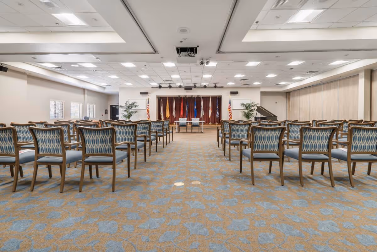 A large conference or meeting room with rows of wooden chairs featuring blue patterned upholstery. At the front of the room is a stage with a table and two chairs, flanked by several flags and two potted plants. The room has a patterned carpet and a ceiling with recessed lighting and a projector.
