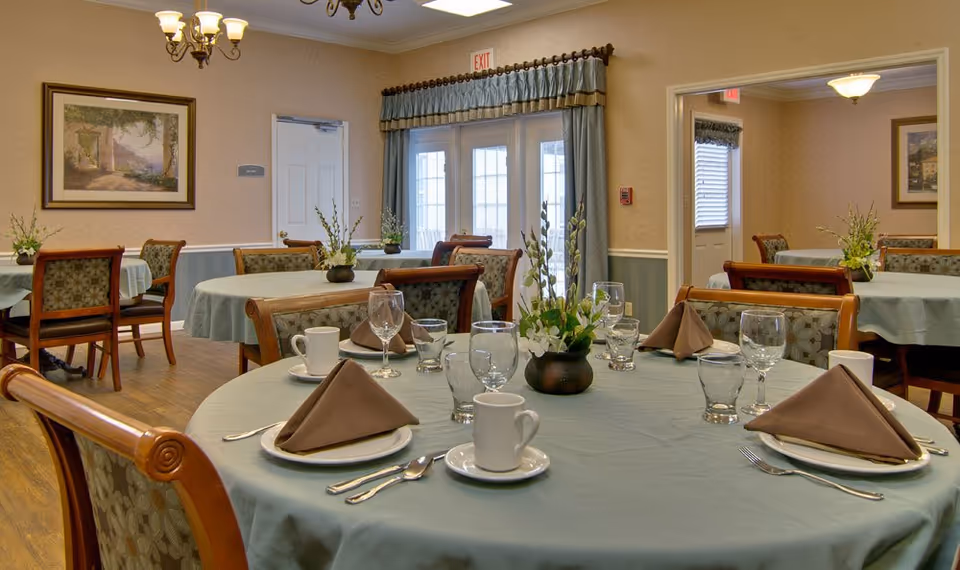 Bright dining room with round tables set with blue tablecloths, brown napkins, glassware and floral centerpieces.