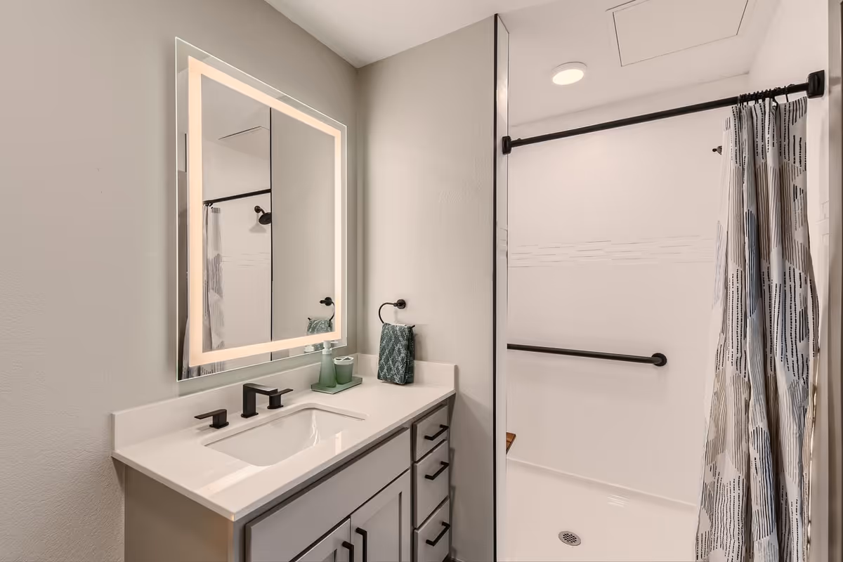 A modern bathroom featuring a white vanity with black handles and a rectangular sink. Above the sink is a large, illuminated mirror with a soft glowing border. To the right is a walk-in shower with a black grab bar, a black showerhead, and a patterned shower curtain. The walls are painted light gray.