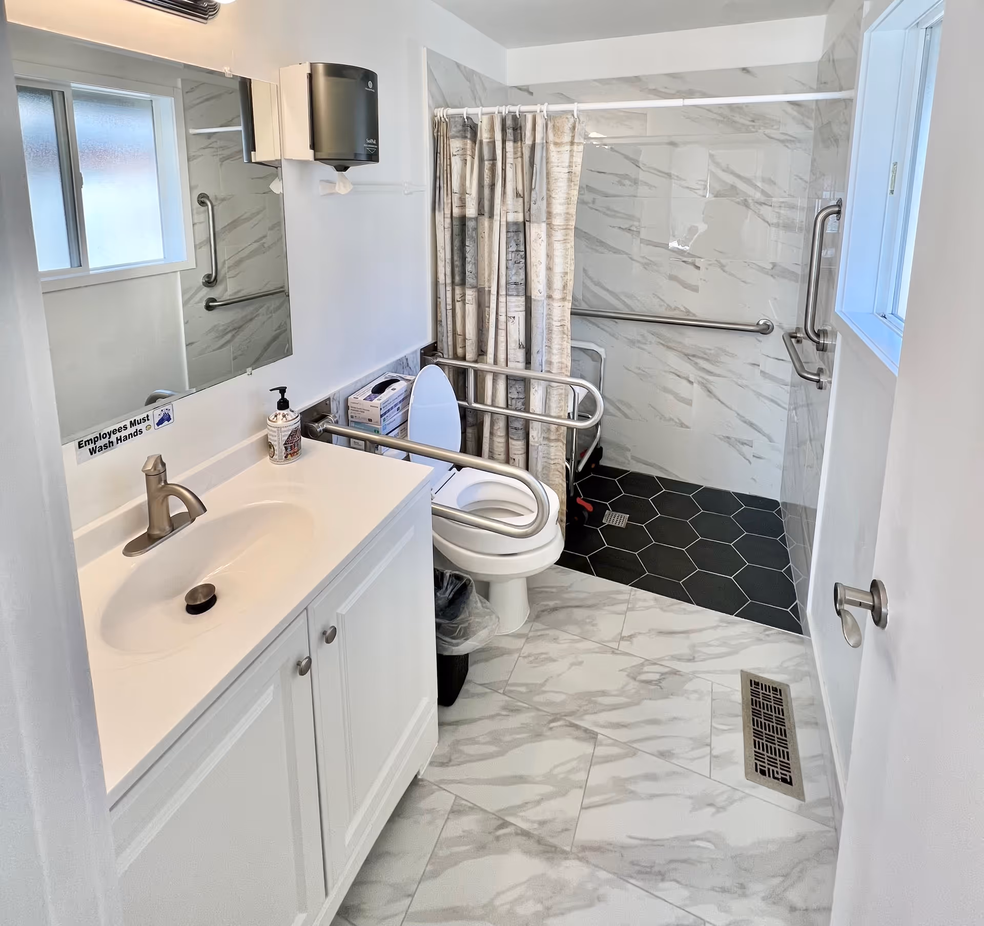 A clean, accessible bathroom with white marble-patterned floor tiles and walls. It features a white sink with a silver faucet, a mirror above it, and a soap dispenser. There is a toilet with safety grab bars on both sides and a shower area with a curtain and black hexagonal floor tiles. The shower has grab bars for accessibility. A window provides natural light, and a sign near the sink reads 'Employees Must Wash Hands.'