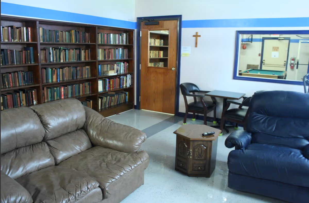 A cozy common area featuring a brown leather couch and a blue leather armchair, a small wooden coffee table with a remote control on it, a wooden bookshelf filled with books, a wooden door with a glass panel, a small table with two chairs, a wall-mounted wooden cross, and a large mirror reflecting a pool table and other room details.