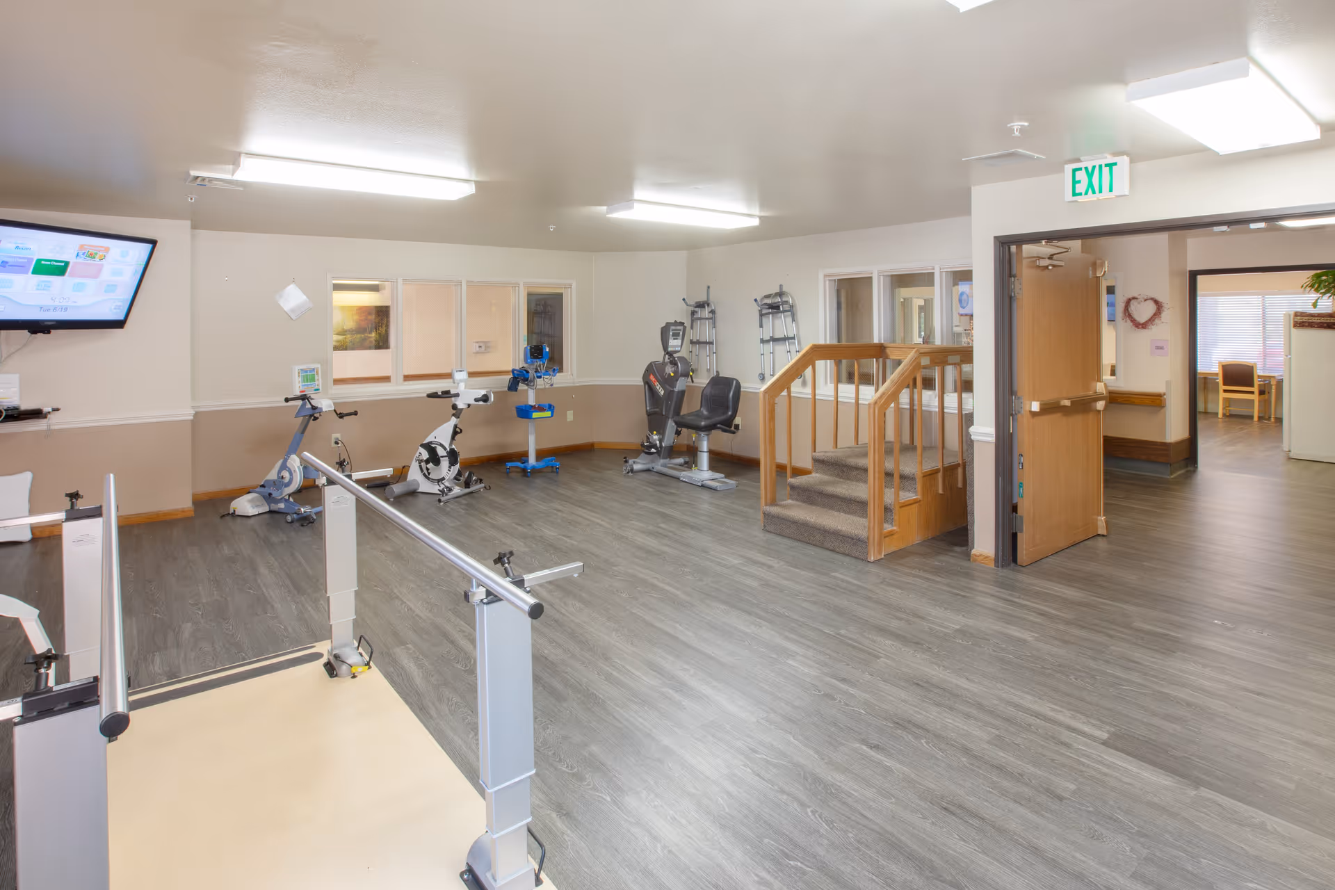 A rehabilitation or physical therapy room in a senior care facility with exercise equipment including stationary bikes and parallel bars for walking practice. The room has wood-look flooring, beige and white walls, a TV mounted on the wall, and an exit door leading to another area of the facility.