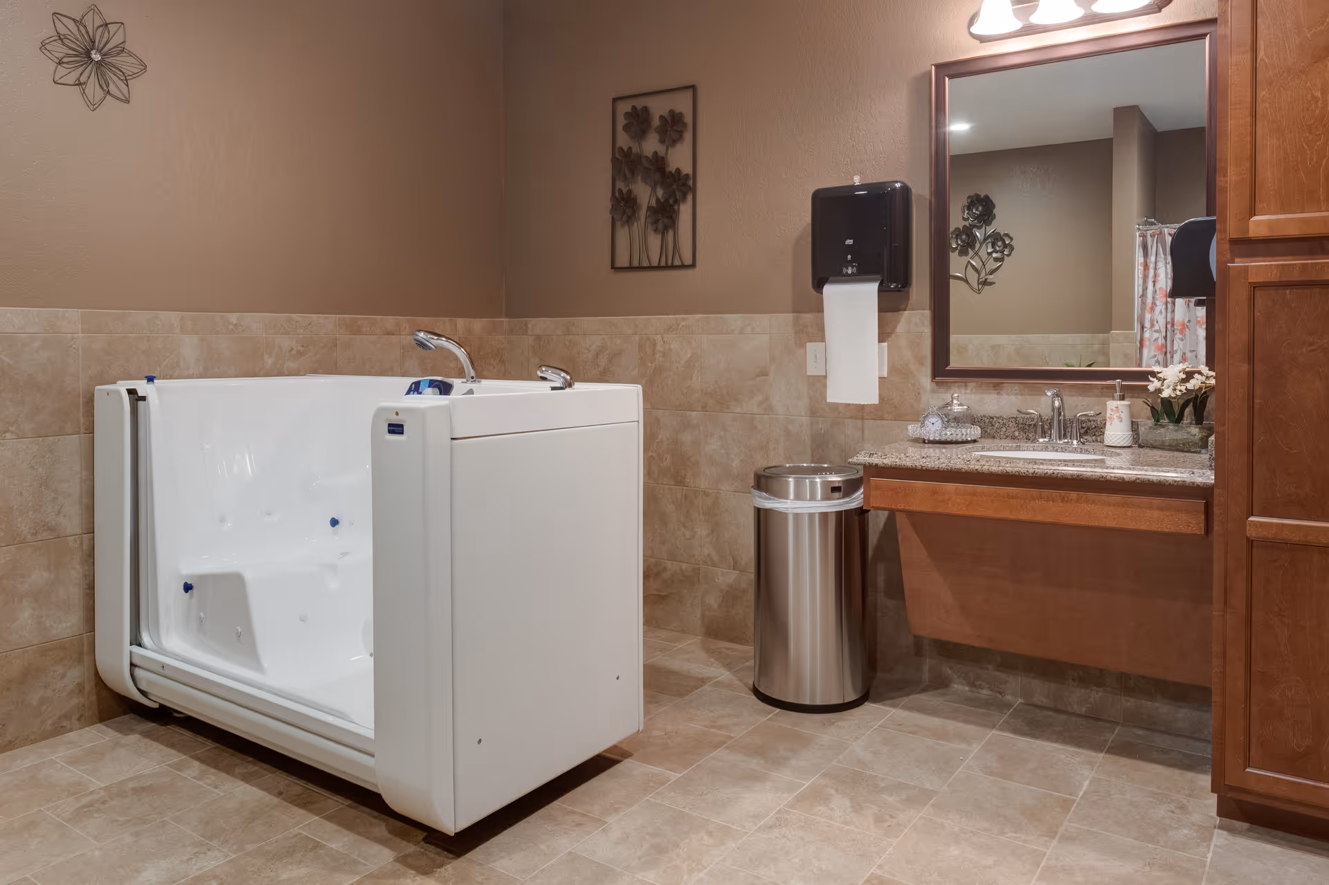 A bathroom with beige tiled walls and floor, featuring a white walk-in bathtub with built-in seat and grab bars. To the right, there is a wooden vanity with a granite countertop, a sink, a large mirror above it, a stainless steel trash can, and a wall-mounted paper towel dispenser. Decorative metal wall art is visible on the walls.