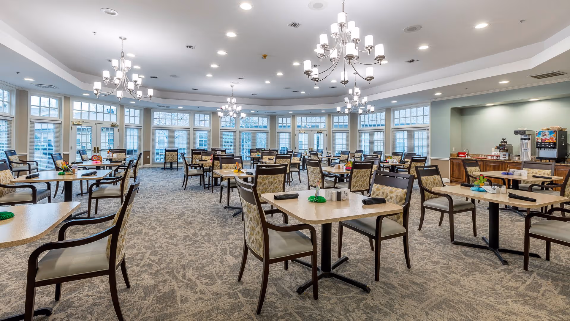 A spacious dining room with multiple tables and chairs arranged neatly. The room features large windows allowing natural light to fill the space, elegant chandeliers hanging from the ceiling, and a beverage station with a coffee machine and drink dispenser in the corner.