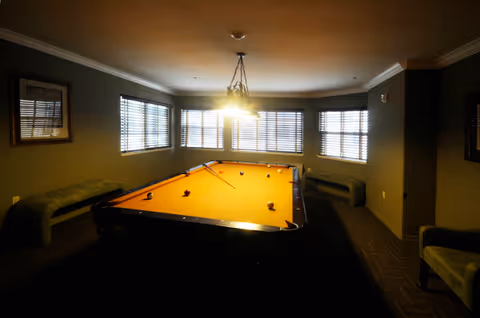 A dimly lit room with a pool table in the center, surrounded by windows with blinds. There are benches along the walls and a hanging light fixture above the pool table.