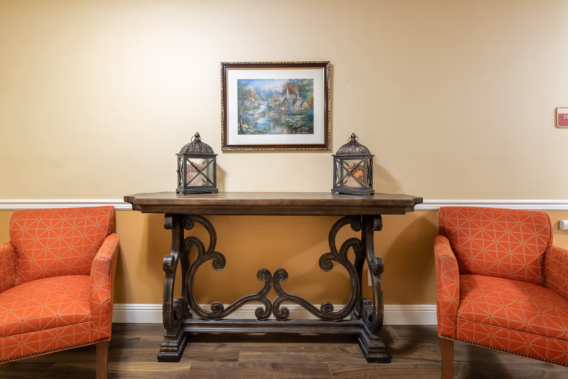Two patterned orange chairs flank a decorative wooden console table topped with lanterns and a framed landscape painting on the wall.