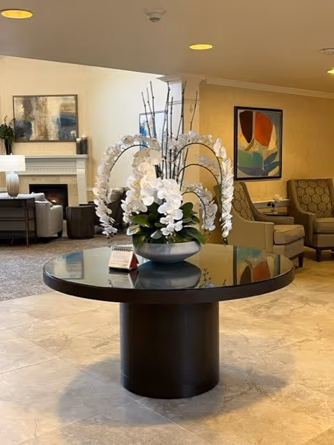 A round black table with a large floral arrangement of white orchids in a silver bowl sits in the center of a senior living facility common area. In the background, there are comfortable armchairs, a fireplace, and framed abstract artwork on the walls.