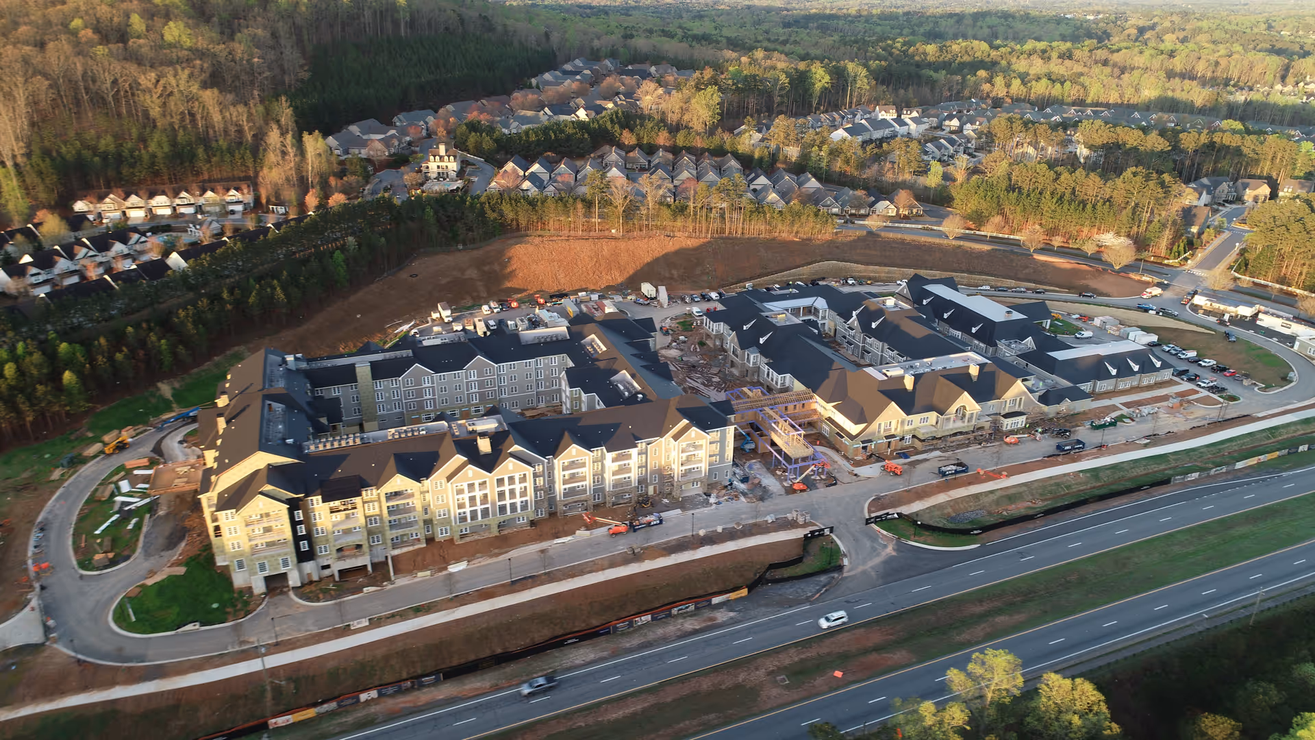 Aerial view of a large senior living complex under construction beside a highway, surrounded by trees and nearby houses.