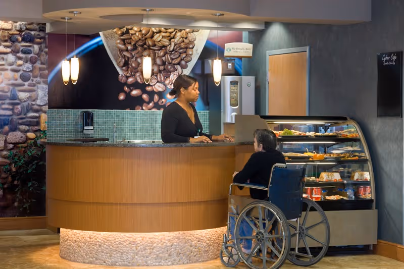 A woman behind a curved wooden counter with a stone base is assisting a person in a wheelchair at a coffee shop or cafe area inside a facility. Behind the counter, there is a large image of coffee beans and a coffee machine. To the right, there is a display case with various food items.