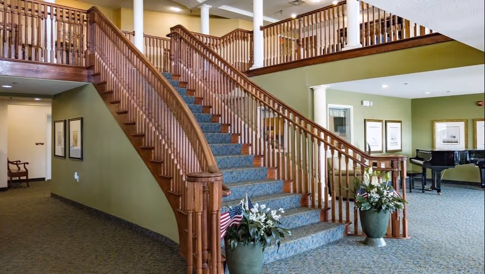 Spacious lobby featuring a curved wooden staircase with carpeted steps, potted plants, and a grand piano.