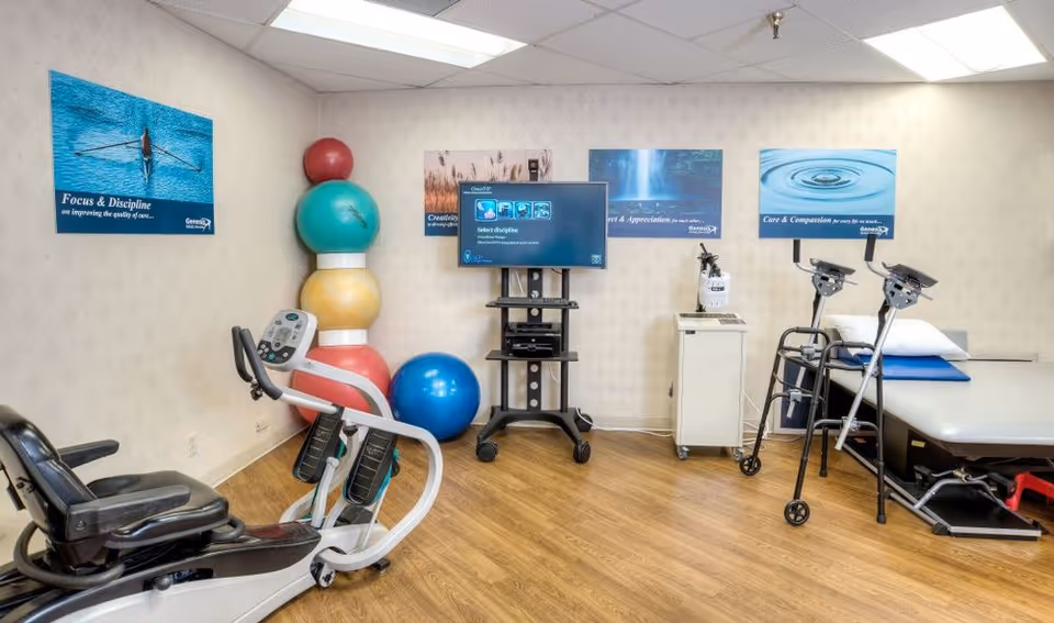 Physical therapy/exercise room with a recumbent bike, stacked therapy balls, a TV on a cart, walkers, and a treatment table.