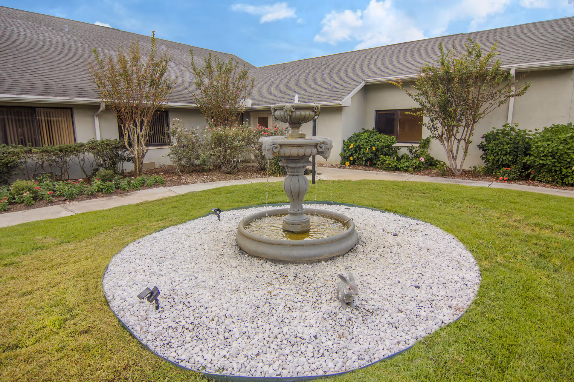 Outdoor courtyard area at Lake Port Square featuring a central stone fountain surrounded by white gravel and green grass, with shrubs and small trees planted along the building walls under a partly cloudy sky.