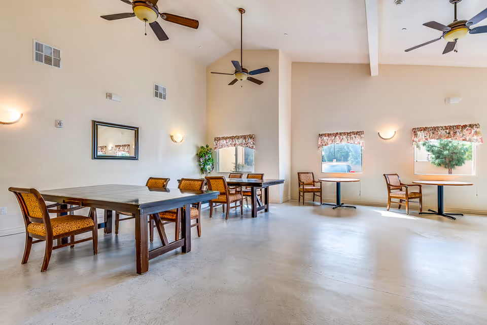 A spacious senior living facility dining room with multiple wooden tables and chairs arranged neatly. The room has high ceilings with ceiling fans, large windows with floral valances allowing natural light to fill the space, and light-colored walls with wall-mounted lights and a mirror.