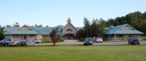 Front view of a single-story senior living facility with green metal roofs, a central peaked entrance, parked cars, and a grassy lawn.
