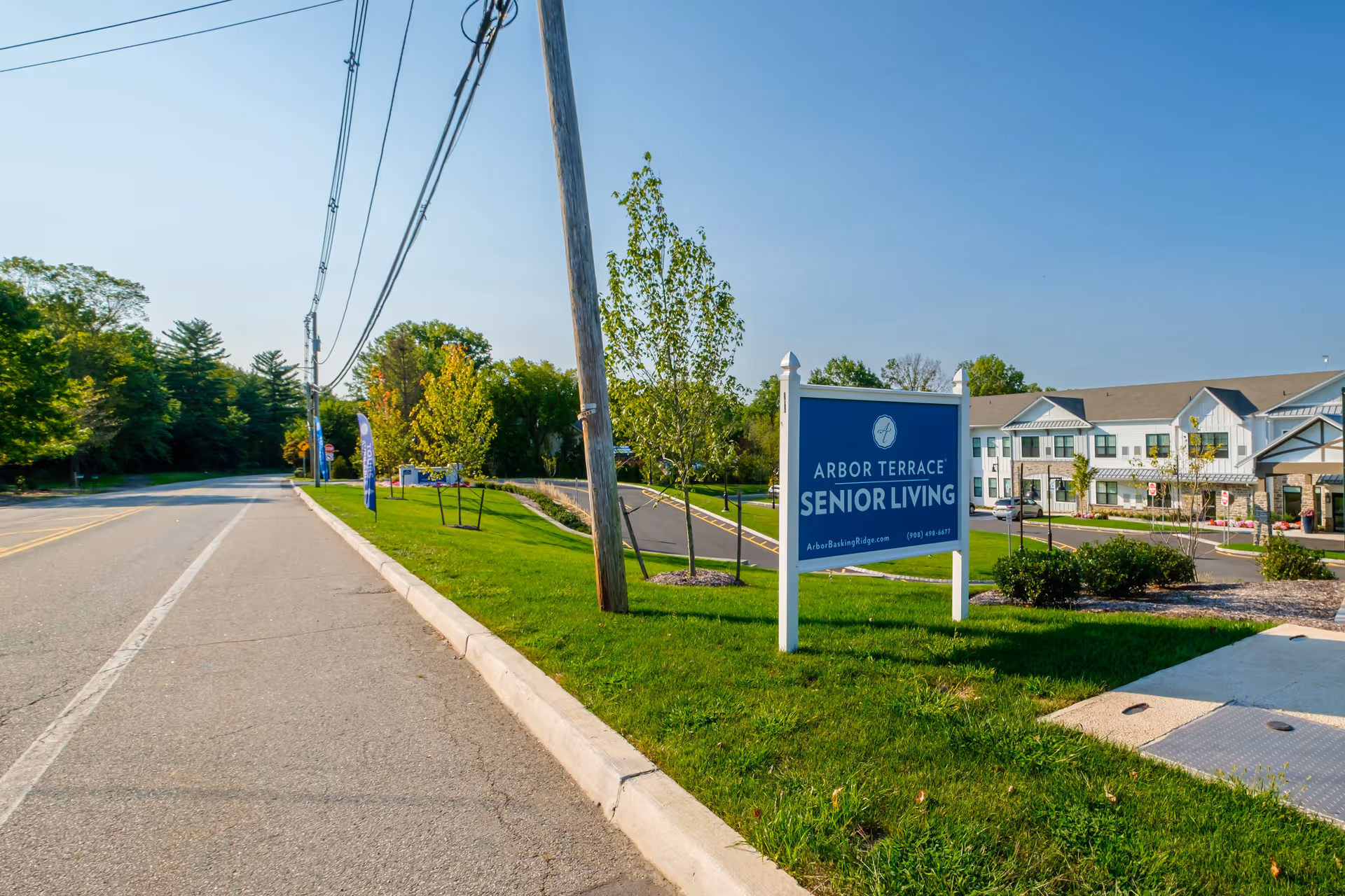 View of the entrance to Arbor Terrace Basking Ridge senior living facility with a blue sign displaying the facility name and contact information, a paved road, green grass, young trees, and a two-story building in the background under a clear blue sky.