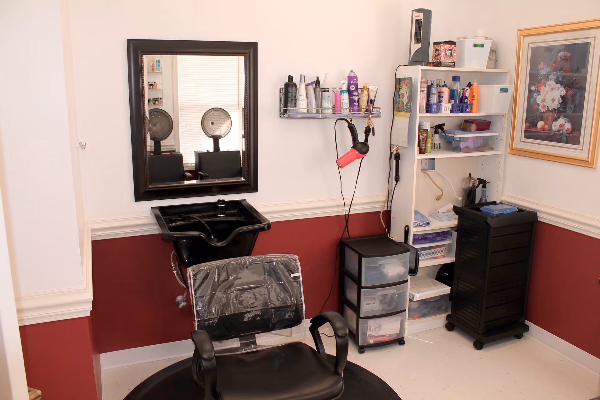 Interior of a hair salon area with a black salon chair covered in plastic, a black hair washing sink, a large mirror on the wall, a shelf with various hair care products, a hair dryer hanging on the wall, and storage drawers and shelves filled with more hair care supplies. There is a framed floral painting on the wall and the lower half of the walls are painted dark red with white trim.