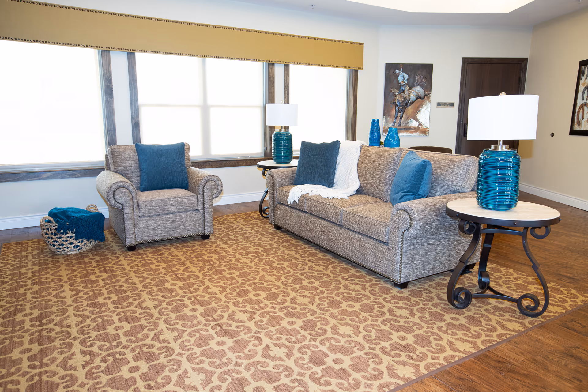 A cozy living room area with a patterned brown and beige rug, a gray upholstered armchair and matching sofa with blue cushions, two round side tables with blue ceramic lamps, a basket with a blue blanket, and large windows with yellow valances letting in natural light.