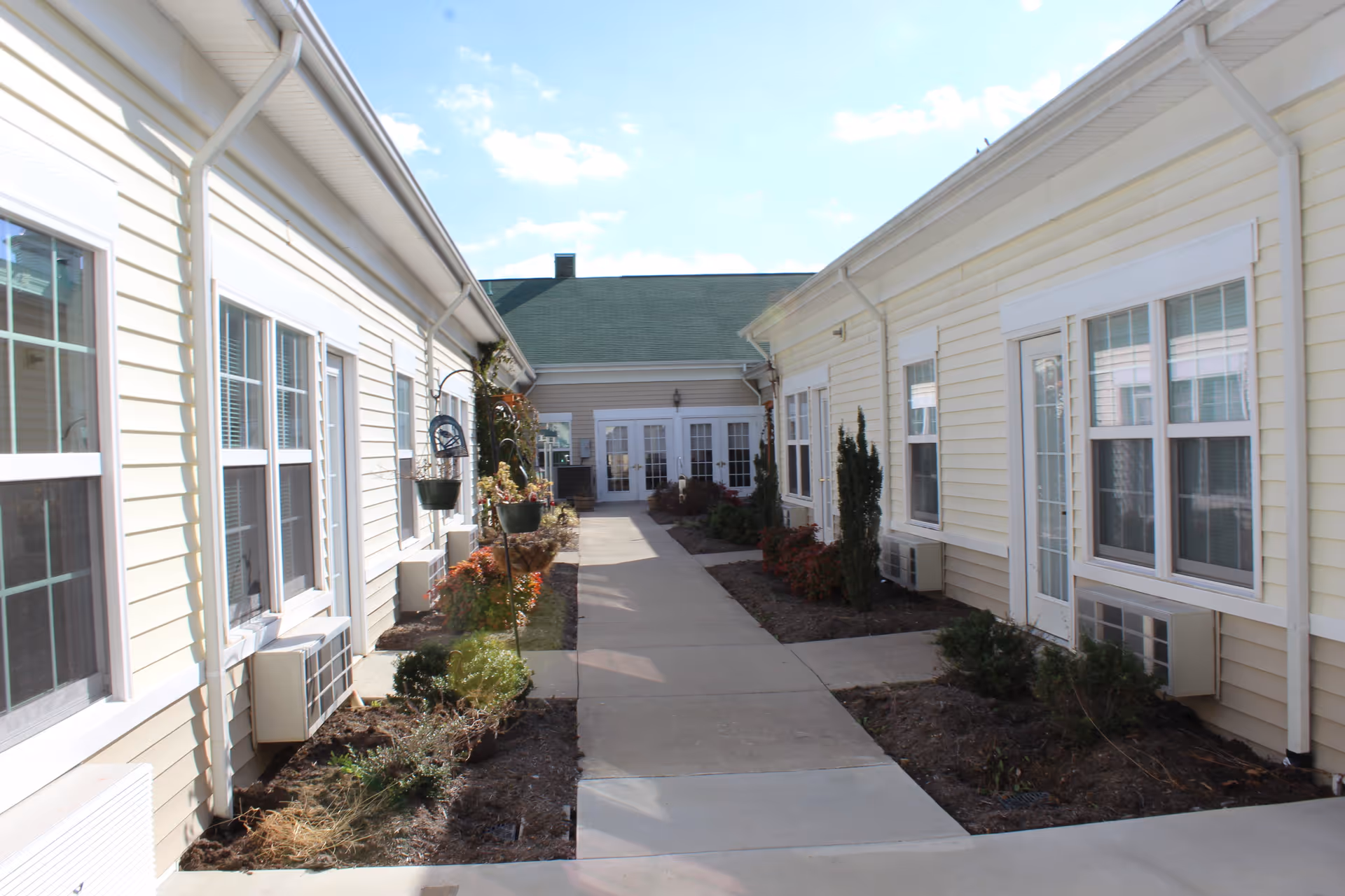Sunlit outdoor courtyard walkway between two single-story yellow-sided building wings with windows, planters, and a path leading to a central entrance.