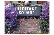 A Heritage Square entrance sign mounted on a brick base surrounded by purple flowers and greenery.