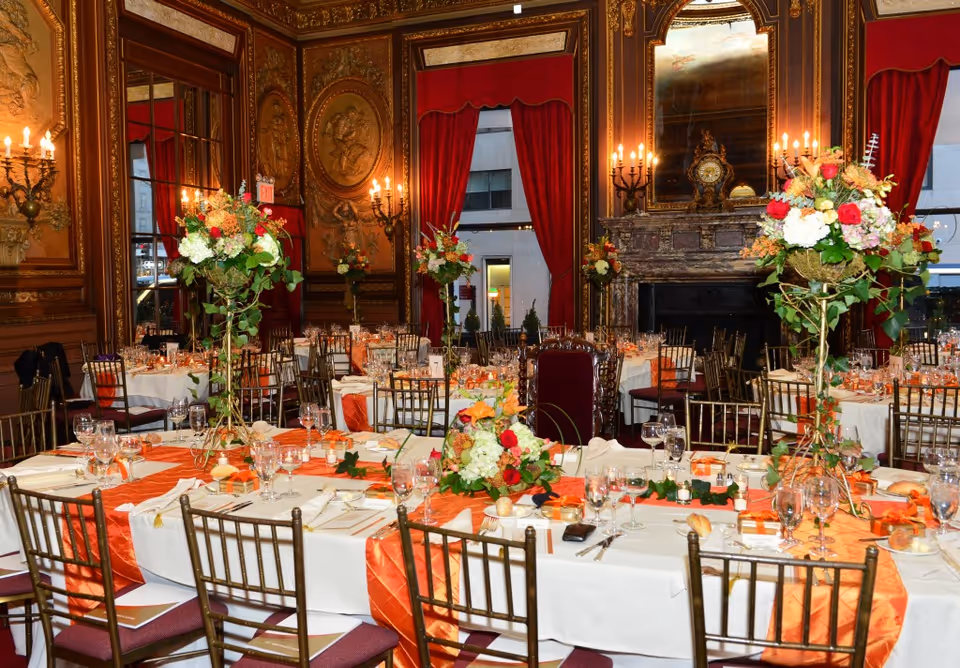 Ornately decorated banquet hall with tables set for a formal dinner, orange runners, tall floral centerpieces, gilded walls, and a fireplace.