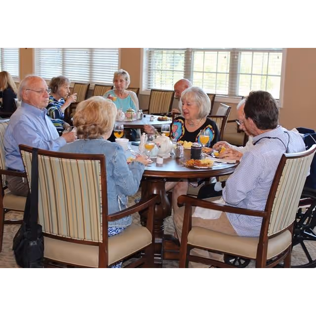 A group of elderly people sitting around a round dining table enjoying a meal together in a well-lit room with large windows and beige walls.