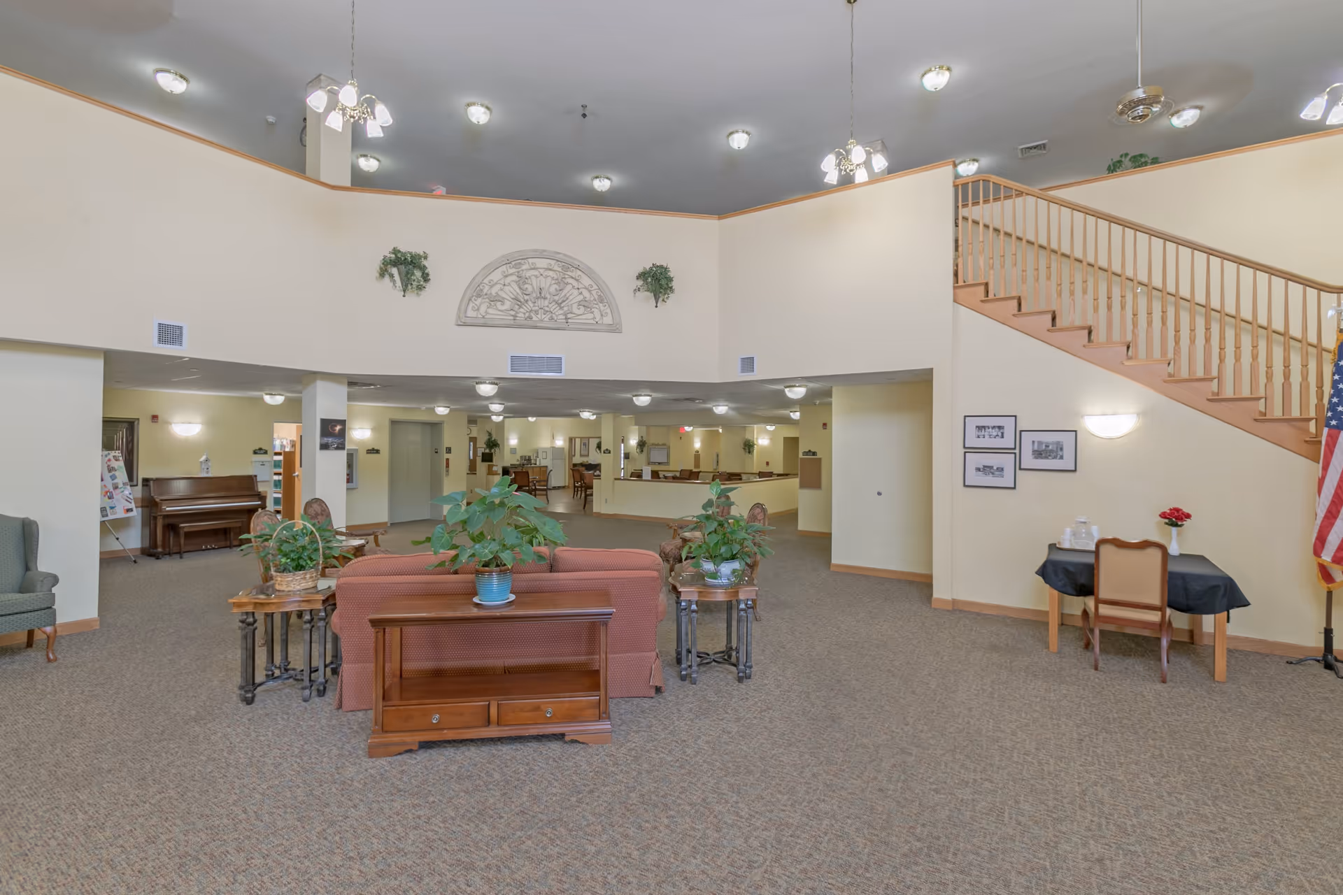 A spacious senior living facility common area with a high ceiling and multiple ceiling lights. The room features a red sofa with potted plants on side tables, a wooden piano in the background, a staircase with wooden railings on the right, and a small table with a chair and an American flag beside it. The walls are light-colored with decorative plants and framed pictures.