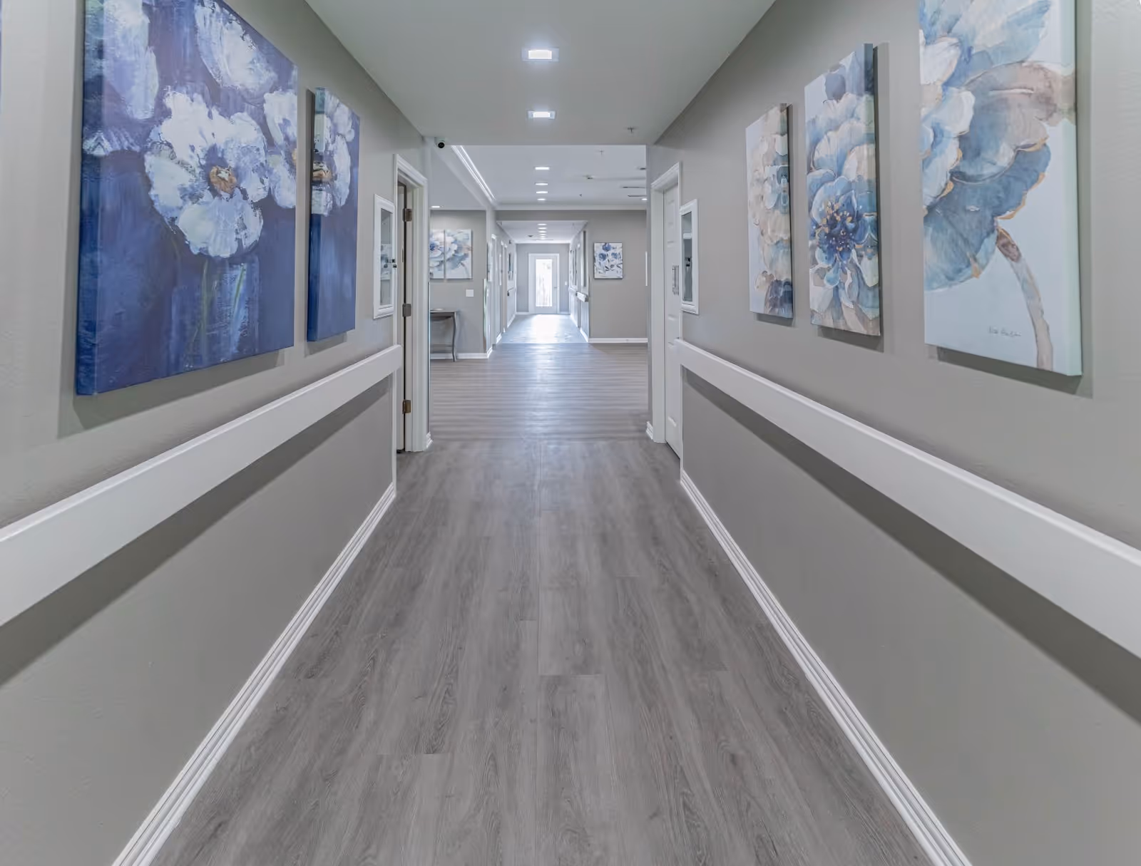 A clean, well-lit hallway in an assisted living facility with light gray walls and wood flooring. The hallway is decorated with large floral paintings on both sides and has white handrails along the walls. Several doorways line the corridor, and recessed ceiling lights illuminate the space.