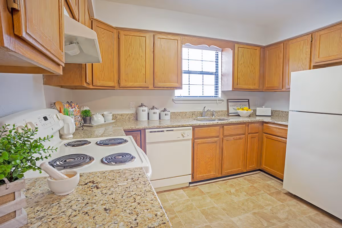 A bright kitchen with wooden cabinets, a white electric stove, a white refrigerator, a dishwasher, and a window above the sink. The countertops are granite with various kitchen items including a plant, a mortar and pestle, canisters, and a bowl of lemons.