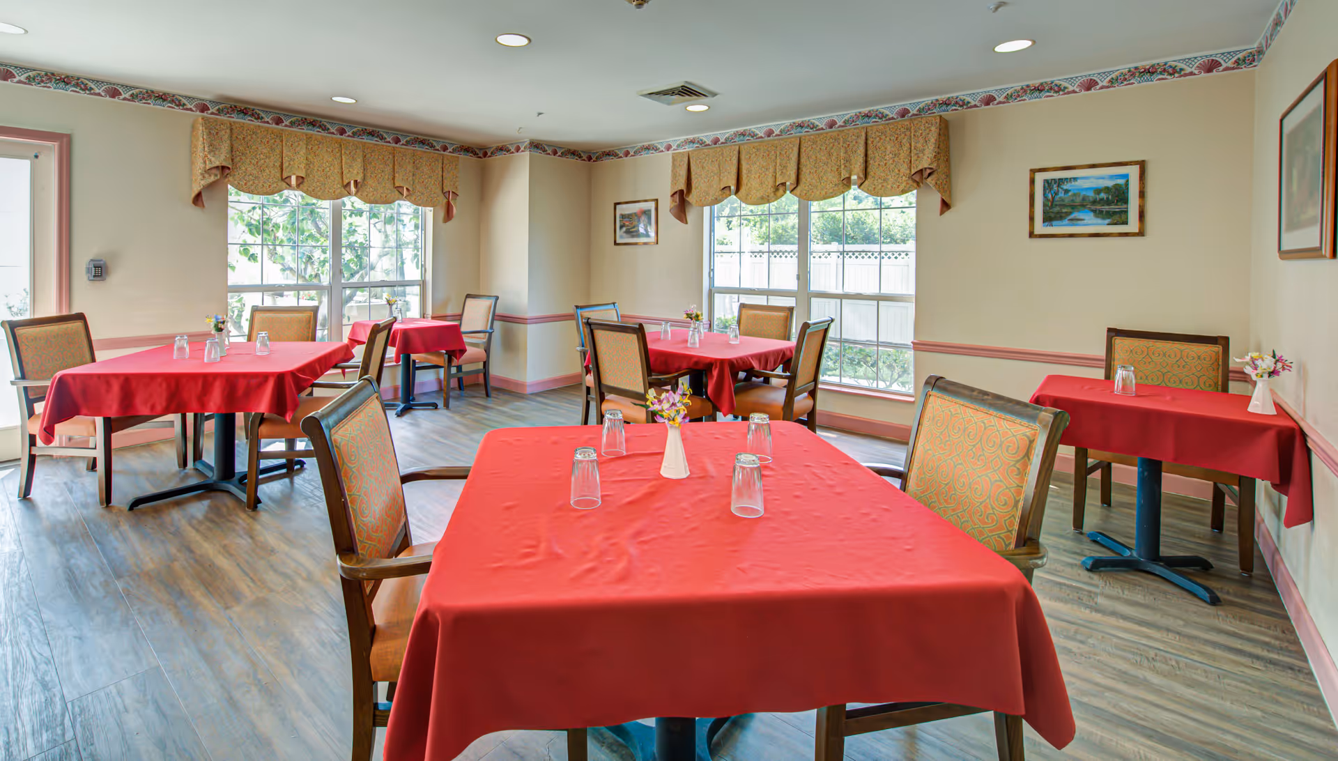 A dining room with several tables covered in red tablecloths, each set with upside-down drinking glasses and small flower vases. The room has large windows with beige valances, framed paintings on the walls, and wooden chairs with patterned upholstery. The floor is wood-style laminate.
