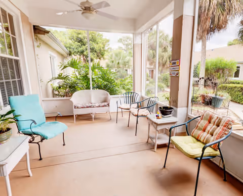 A covered outdoor patio area with several metal chairs featuring colorful cushions, a small white side table with a potted plant, and lush green plants visible outside the screened enclosure.