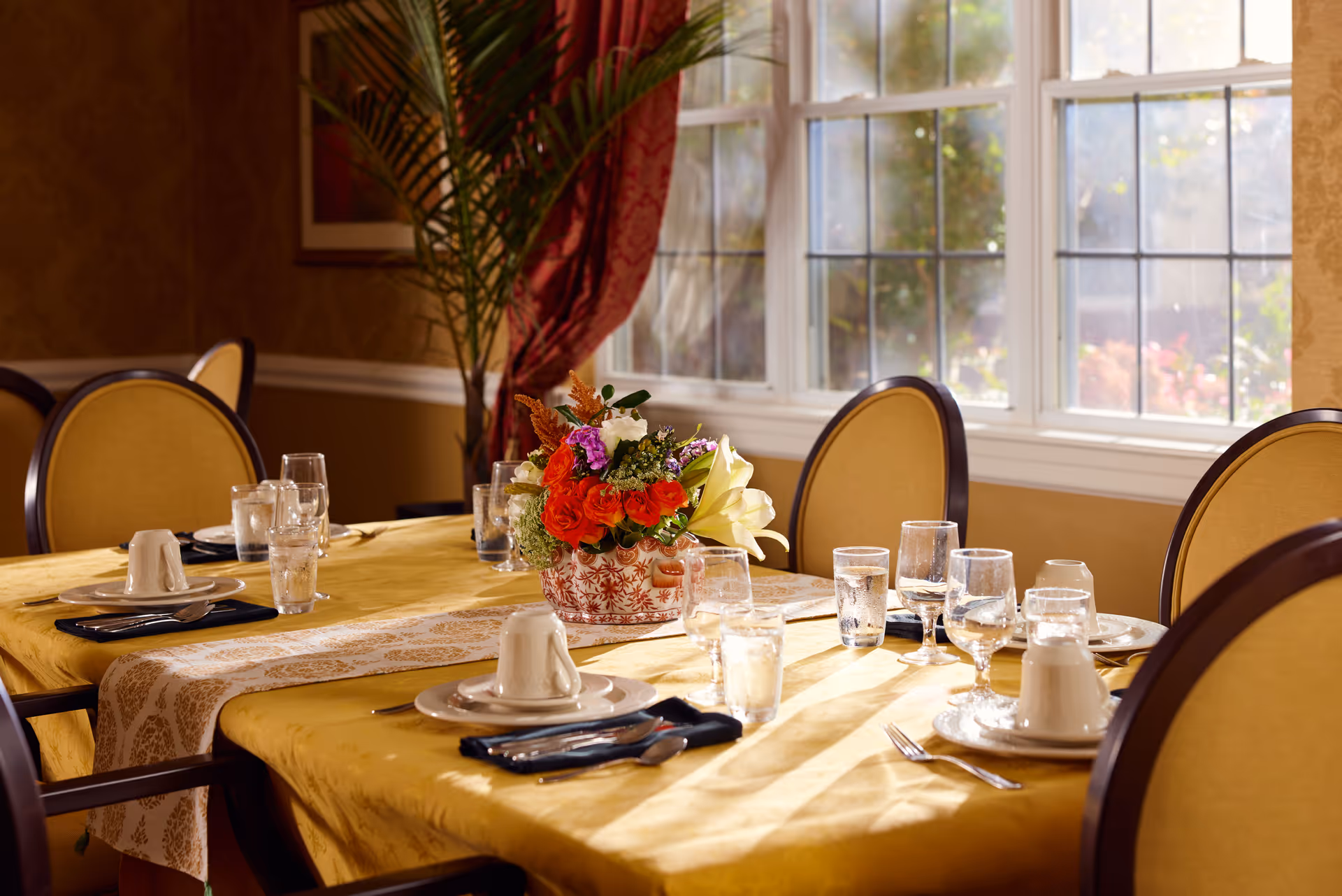 A dining table set with plates, cups, utensils, and glasses filled with water. A floral centerpiece with red, white, and purple flowers is placed in the middle of the table. The room has large windows with sunlight streaming in, yellow upholstered chairs, and warm-toned walls with a plant and red curtains in the background.