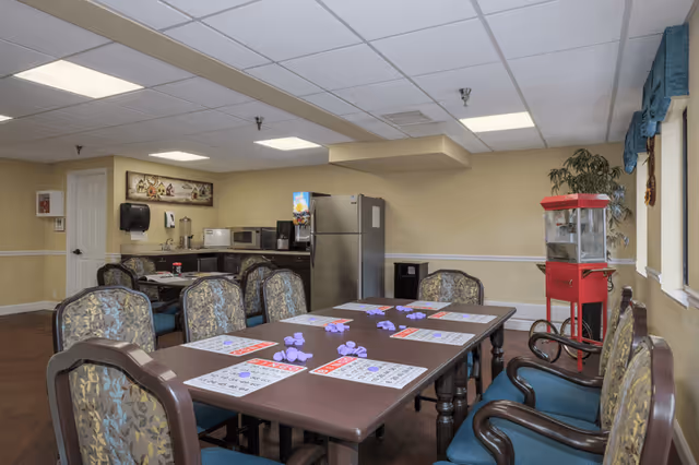 Communal activity/dining room with a long table set for bingo, upholstered chairs, a kitchenette and a popcorn machine.