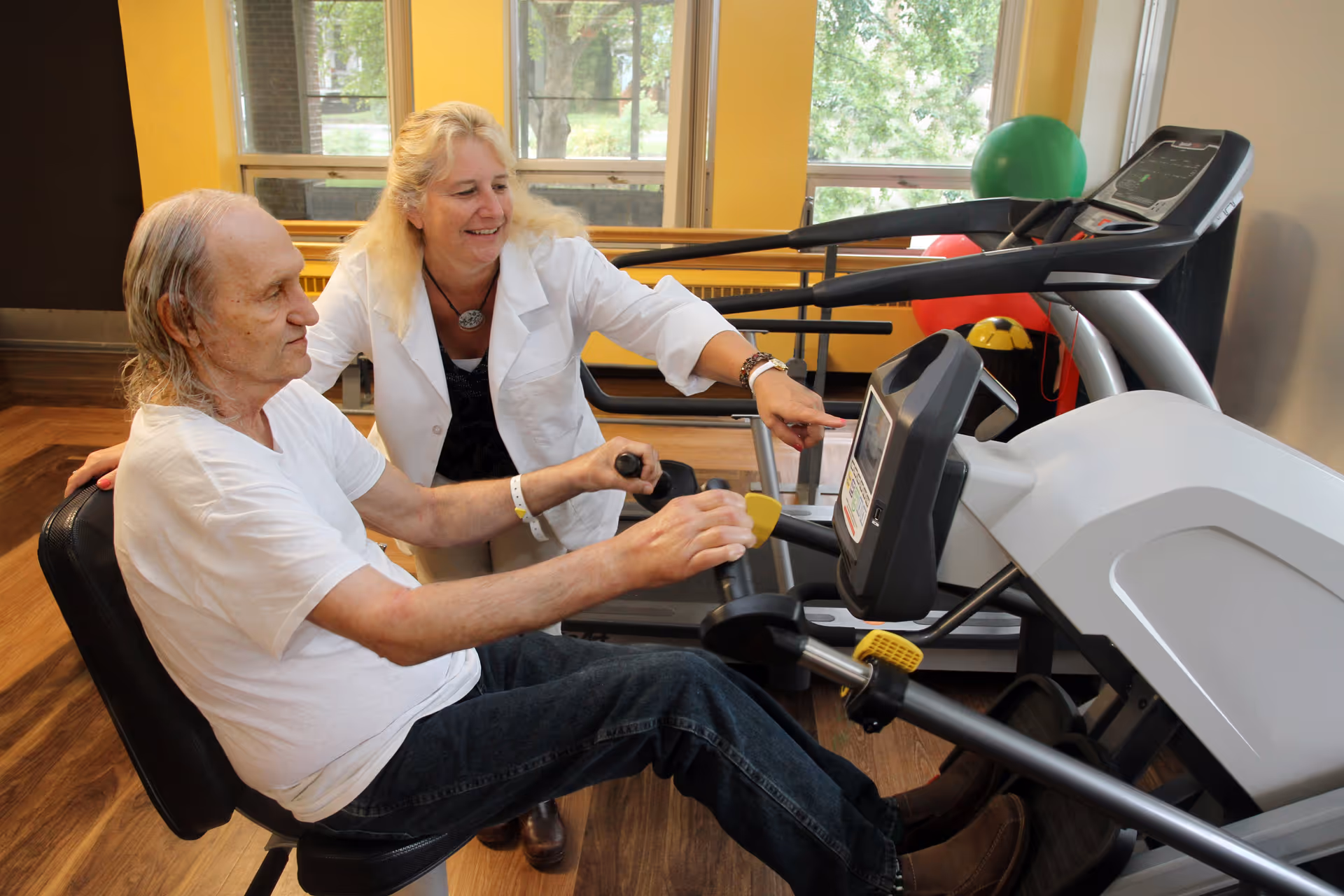 An elderly man using a seated exercise machine in a rehabilitation center while a female therapist or instructor in a white coat assists and points at the machine's display. The room has large windows with a view of trees outside and exercise equipment in the background.