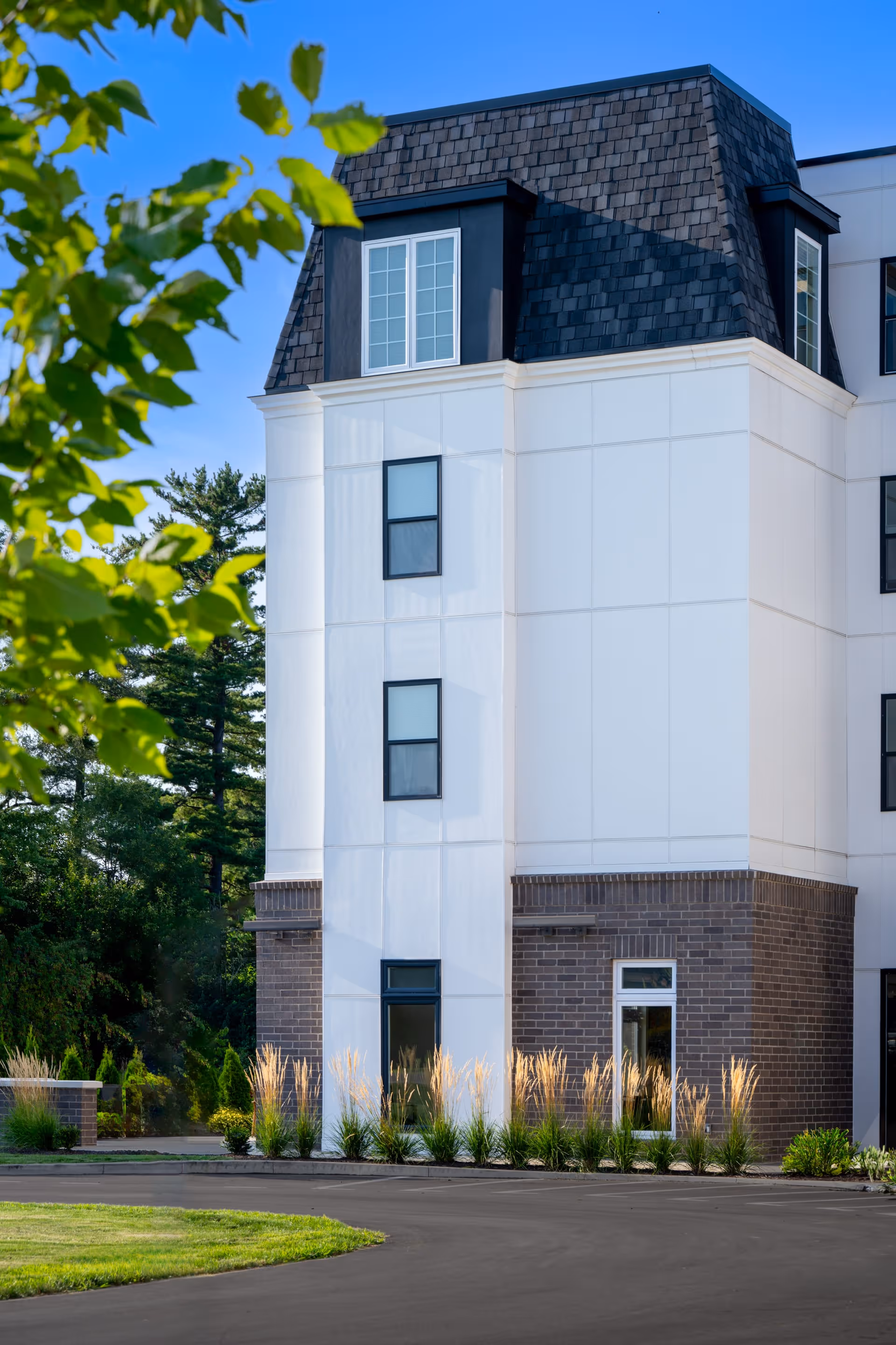 Exterior front corner of a multi-story senior living building with white and brick facade, windows, landscaping, and a shingled roof against a blue sky.
