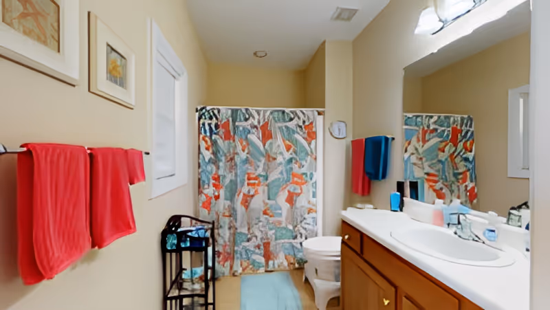 A bathroom with beige walls featuring a shower with a colorful abstract patterned curtain, a white toilet, a wooden vanity with a white sink, and a large mirror above it. There are red towels hanging on a rack on the left wall and a blue towel hanging near the sink. A small black metal shelf holds toiletries next to the shower.
