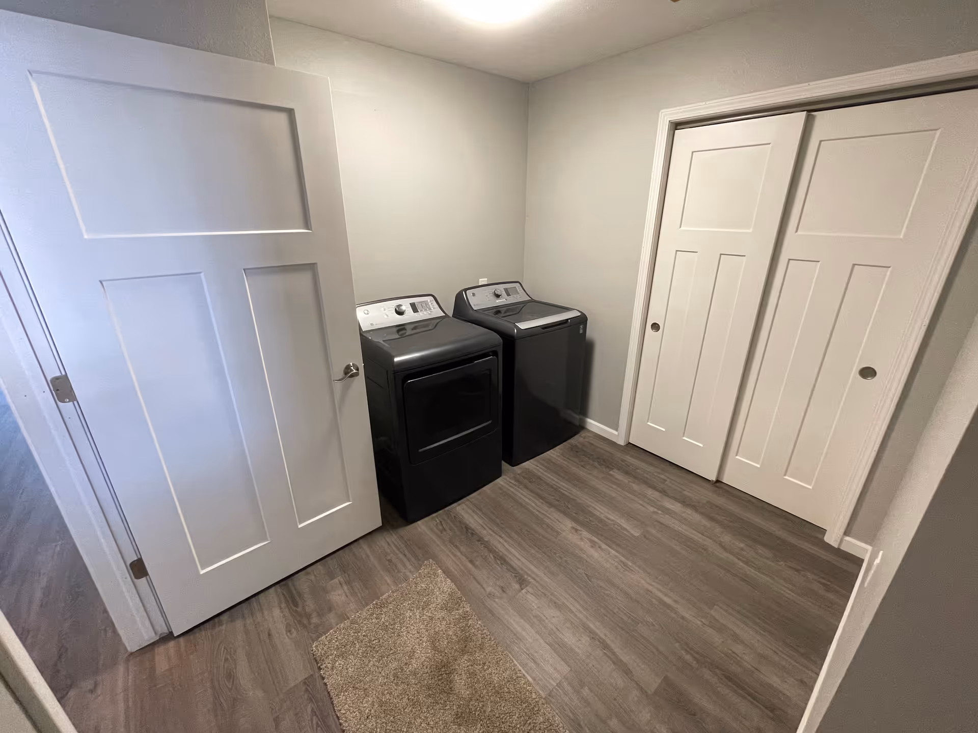 A laundry room with a washer and dryer side by side against a light gray wall. The room has wood-look flooring, a small beige rug in front of the machines, a white door partially open on the left, and white sliding closet doors on the right.