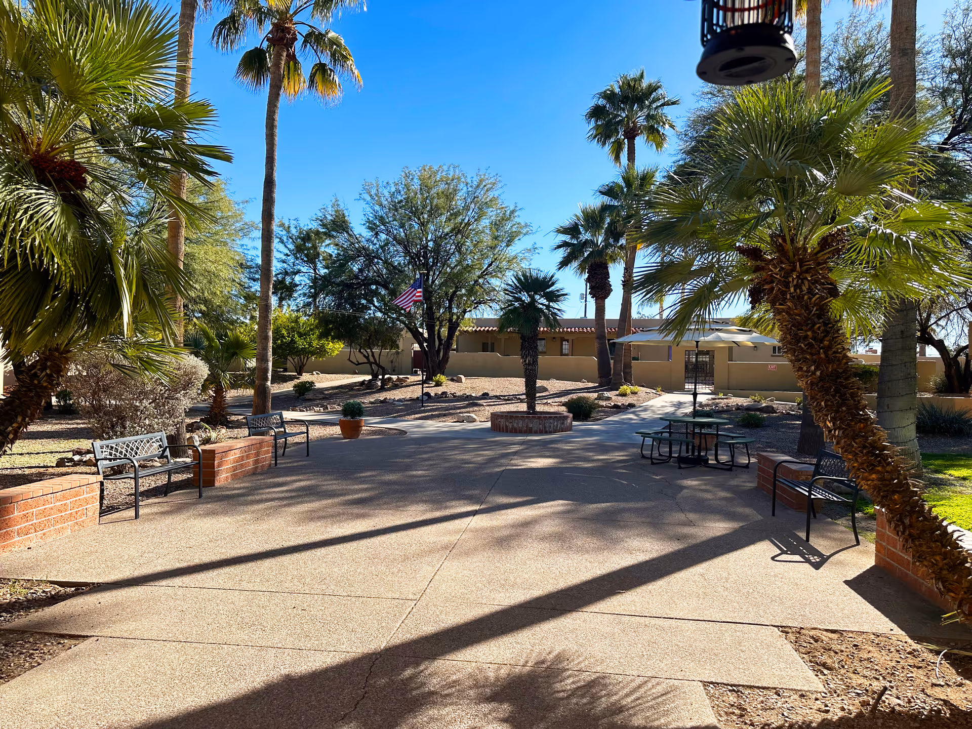 Sunlit outdoor courtyard with palm trees, benches, a picnic table and an American flag.