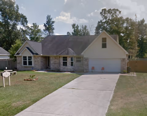 Single-story brick house with a two-car garage, a concrete driveway, and a small front yard with grass and a few plants. There are two orange chairs placed in front of the garage door. Trees are visible in the background under a partly cloudy sky.