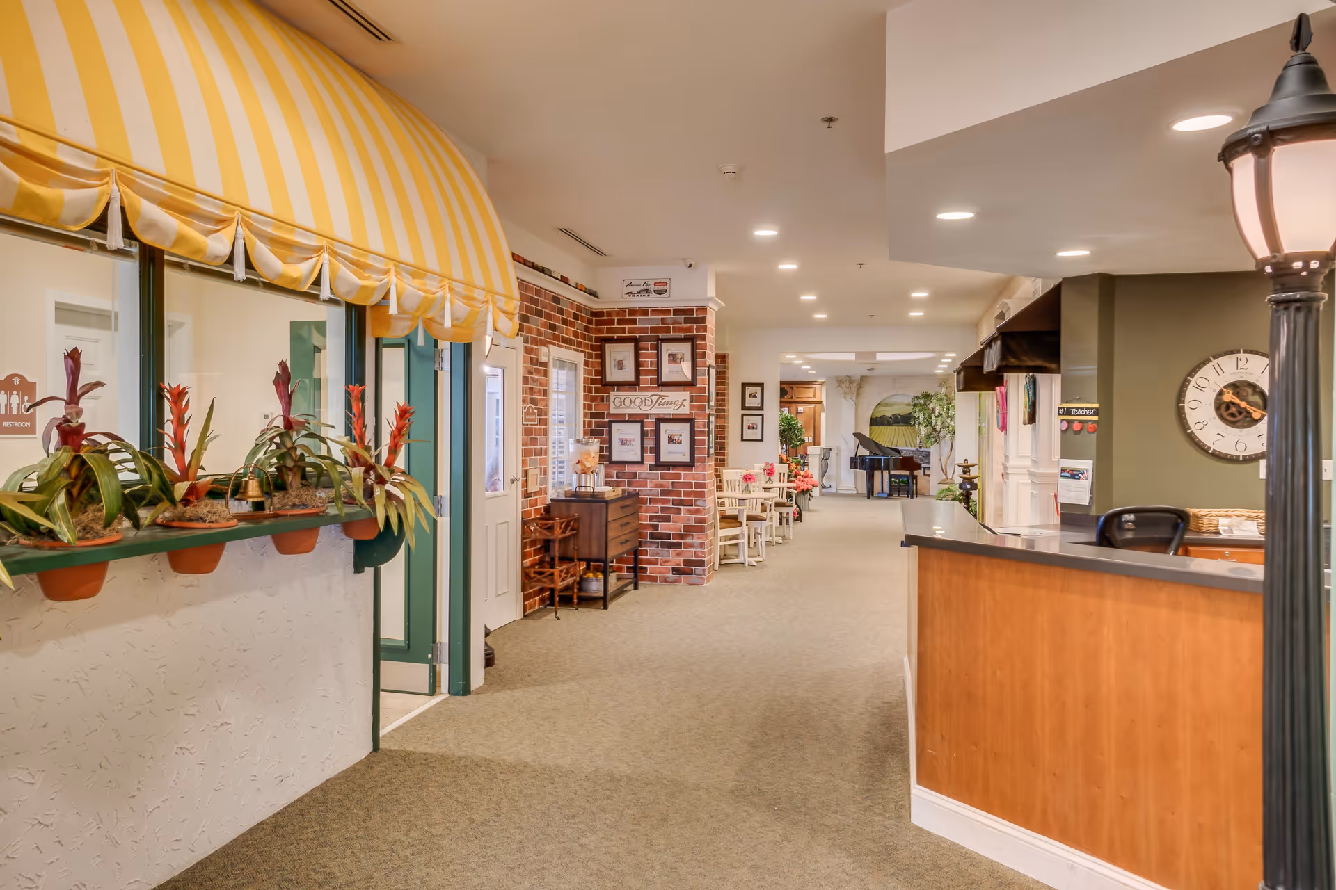 Interior view of a senior living facility hallway with a reception desk on the right, a yellow and white striped awning over a window with potted plants on the left, brick accent wall with framed pictures, small tables and chairs along the hallway, and a piano in the background.