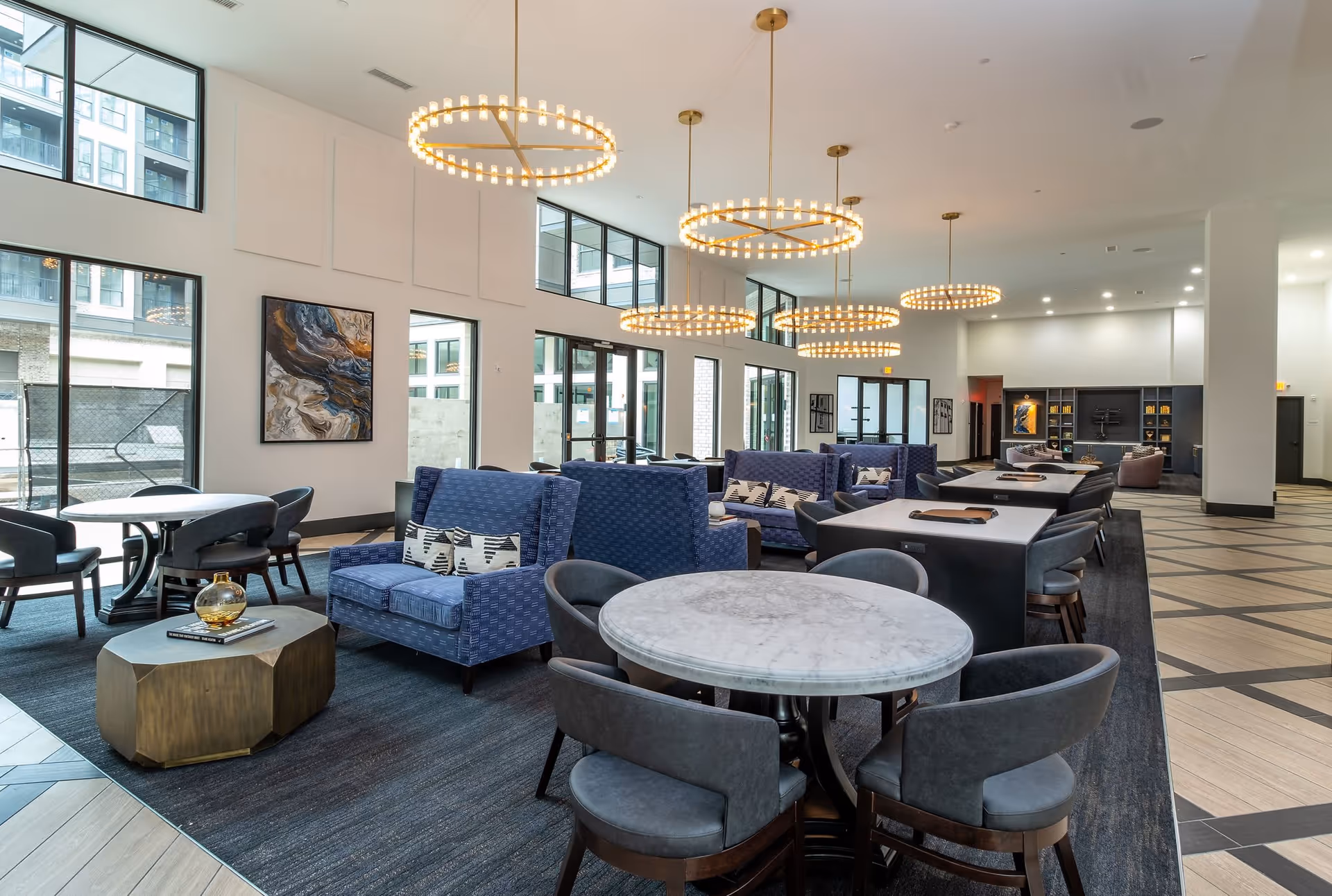 A spacious and modern common area with multiple seating options including blue upholstered sofas, round marble tables with gray chairs, and a geometric coffee table. Large windows allow natural light to fill the room, and several circular chandeliers hang from the ceiling. The space has a contemporary design with artwork on the walls and a patterned floor.